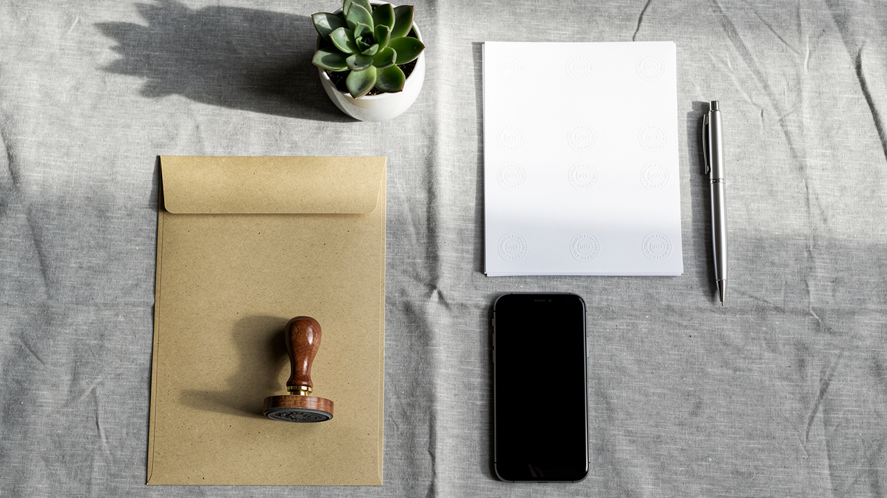 Un plic maro cu o ștampilă de lemn, un smartphone negru, un blocnotes alb și un certificat constatator punct de lucru sunt așezate l&acirc;ngă un stilou și un ghiveci de plante suculente pe o suprafață textilă gri.